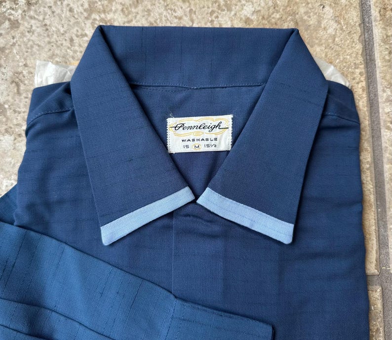 May include: A navy blue collared shirt with light blue trim on the collar and cuffs. The shirt has a label that reads "Pennleigh Washable 15 M 15 1/2". The shirt is folded neatly.