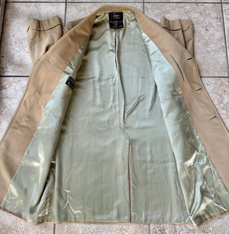 May include: A tan overcoat with a light green satin lining. The coat is open, revealing the interior lining and a label. The coat has buttons and a classic design, suitable for cooler weather. The coat's length is visible.