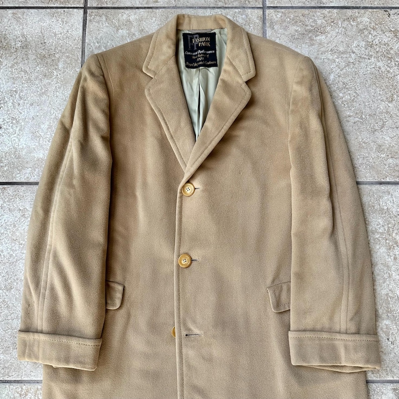 May include: A tan suede coat with a notched lapel, three-button closure, and two front pockets. The coat has a classic design and a soft, textured appearance. The label reads "Fashion Park".