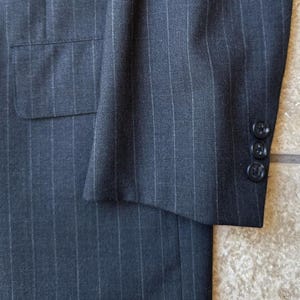 May include: A dark gray pinstripe suit jacket with a single button closure and a pocket on the left side. The jacket has a long sleeve with a button cuff.