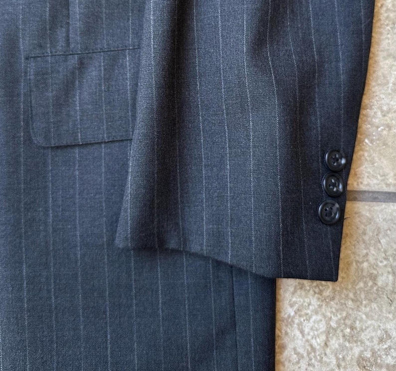 May include: A dark gray pinstripe suit jacket with a single button closure and a pocket on the left side. The jacket has a long sleeve with a button cuff.
