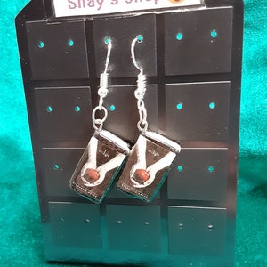 May include: A pair of silver dangle earrings with miniature book charms featuring the cover of the book "Twilight" by Stephenie Meyer. The earrings are displayed on a black earring display card with the text "Shay's shop" at the top.