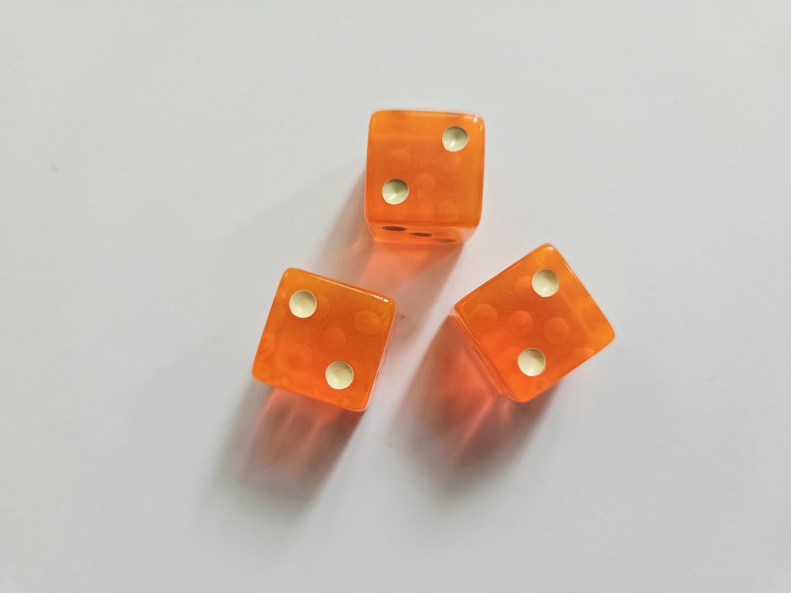 May include: Three translucent orange six-sided dice arranged in a triangular formation. Each die features rounded corners and white circular pips representing the numbers two and four on the visible faces, casting soft shadows on the surface.