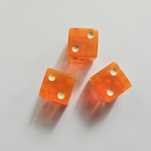 May include: Three translucent orange six-sided dice arranged in a triangular formation. Each die features rounded corners and white circular pips representing the numbers two and four on the visible faces, casting soft shadows on the surface.