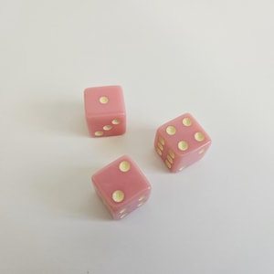 Könnte beinhalten: Drei rosa Würfel mit goldfarbenen Punkten liegen auf einer weißen Oberfläche. Jeder Würfel hat eine unterschiedliche Anzahl sichtbarer Punkte, von eins bis vier. Die Würfel haben eine weiche, pastellrosa Farbe.