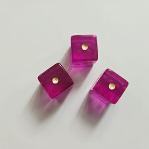 May include: Three translucent magenta six-sided dice arranged in a triangular formation. Each die is constructed from a clear, vibrant pink material and features a single gold-colored circular pip on the top face, indicating the number one.