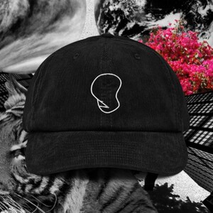 May include: A black corduroy baseball cap featuring a white embroidered outline of a stylised, abstract head shape on the front panel. The cap has a structured crown, a curved brim, and a matching fabric strap with a metal buckle at the back.