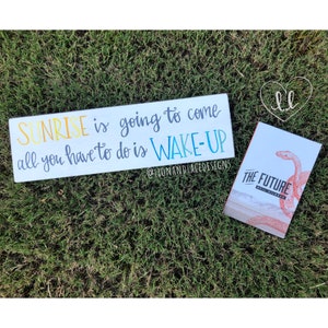 May include: A white wooden sign with the text "SUNRISE is going to come all you have to do is WAKE-UP" in colorful lettering. The sign is on a green grassy surface. A small book with the text "THE FUTURE" is also on the grass.