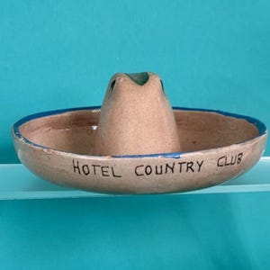 May include: A ceramic ashtray shaped like a sombrero, with a beige base and a blue rim. The words "HOTEL COUNTRY CLUB" are written in black around the edge. The sombrero's crown is in the centre.