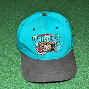 Op de afbeelding: Een blauwgroene honkbalpet met een zwarte klep en een zwarte knoop aan de bovenkant. De voorkant is voorzien van een geborduurd Vancouver Grizzlies-logo, met een bruine beer die een basketbal vasthoudt, en de tekst Vancouver Grizzlies in blauwgroene en witte letters.