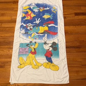 May include: White rectangular beach towel featuring Disney characters Mickey Mouse, Pluto, and Goofy. The top section depicts Goofy snorkeling with colorful fish. The bottom section shows Mickey and Pluto with the text "DO NOT FEED THE FISH!"