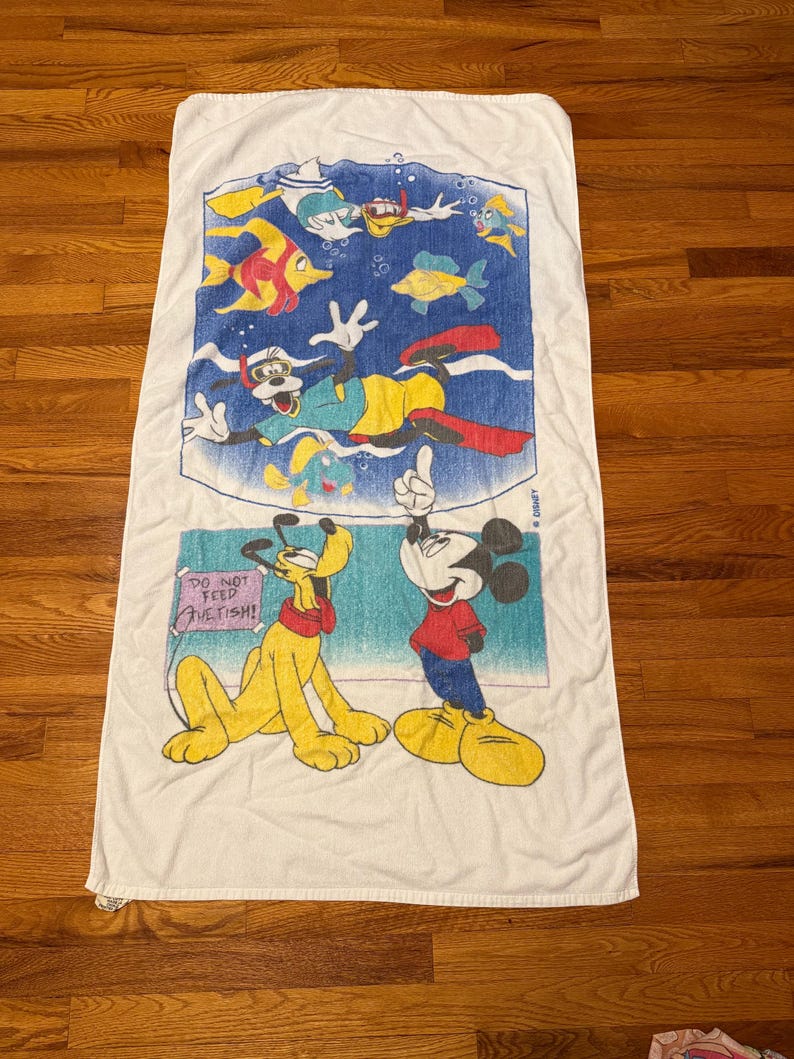 May include: White rectangular beach towel featuring Disney characters Mickey Mouse, Pluto, and Goofy. The top section depicts Goofy snorkeling with colorful fish. The bottom section shows Mickey and Pluto with the text "DO NOT FEED THE FISH!"