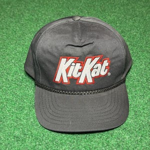 May include: A black baseball cap with the Kit Kat logo embroidered in red and white. The cap has a curved brim and a braided rope detail along the edge. The background is green.