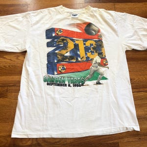May include: White t-shirt featuring a colourful graphic of a baseball player hitting a home run. The graphic includes the text "213", "Camden Yards", and "September 6, 1993".