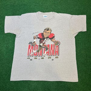 May include: A gray t-shirt with a cartoon image of Joe Montana, the former quarterback for the San Francisco 49ers, wearing a red and white jersey with the number 16. The shirt has the words "Joe Montana 49ers" printed on it.