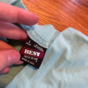 May include: A close-up of a light blue t-shirt collar with a black tag. The tag reads "BEST" and "FRUIT OF THE LOOM" with "MADE IN U.S.A." The tag also indicates the shirt is size "L" for adults and is made of 50% cotton and 50% polyester.