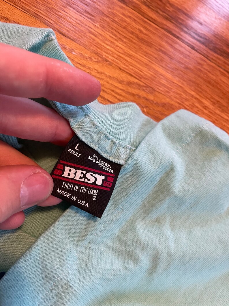 May include: A close-up of a light blue t-shirt collar with a black tag. The tag reads "BEST" and "FRUIT OF THE LOOM" with "MADE IN U.S.A." The tag also indicates the shirt is size "L" for adults and is made of 50% cotton and 50% polyester.