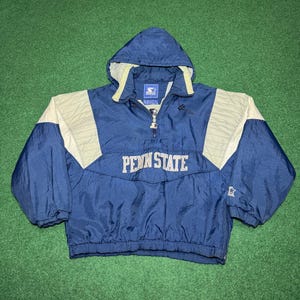 Puede incluir: Chaqueta con capucha azul marino y beige de Penn State con media cremallera. La chaqueta tiene las palabras "PENN STATE" en blanco en el pecho y un pequeño logotipo de Starter en la manga.