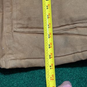 Może przedstawiać: Zbliżenie brązowej kurtki roboczej z kieszenią i miarką. Taśma miernicza pokazuje długość około 81 cm. Kurtka leży na zielonej powierzchni.