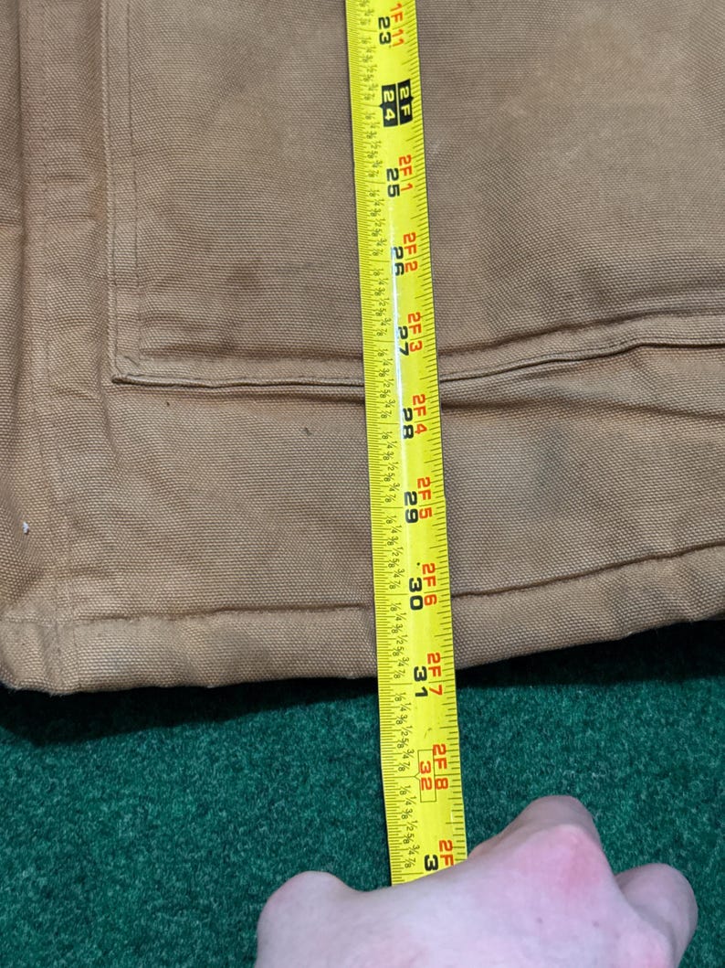 Może przedstawiać: Zbliżenie brązowej kurtki roboczej z kieszenią i miarką. Taśma miernicza pokazuje długość około 81 cm. Kurtka leży na zielonej powierzchni.