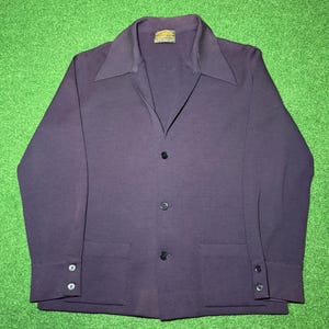 May include: A dark purple blazer with a wide collar, three buttons, and two front pockets. The long-sleeved jacket has buttoned cuffs and a label at the neckline. The blazer is laid flat on a green surface.