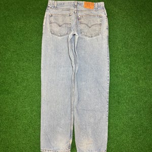 May include: Light blue denim jeans with a classic five-pocket design. The jeans feature a faded wash and a brown leather patch on the back waistband. The back pockets have a signature stitch detail. The jeans are laid flat on a green surface.