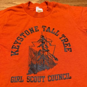 May include: An orange t-shirt with a black graphic and text. The graphic features a stylized tree within a triangle, with the words "KEYSTONE TALL TREE" arched above and "GIRL SCOUT COUNCIL" below.