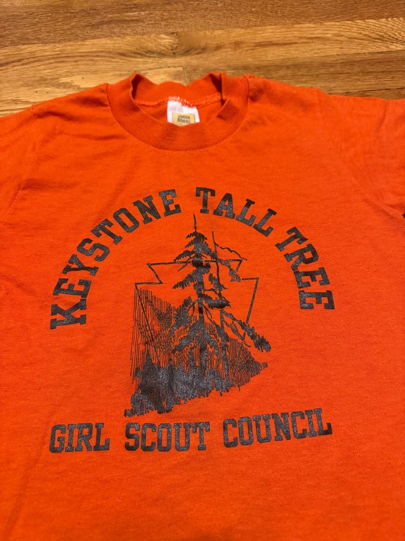 May include: An orange t-shirt with a black graphic and text. The graphic features a stylized tree within a triangle, with the words "KEYSTONE TALL TREE" arched above and "GIRL SCOUT COUNCIL" below.
