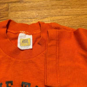 May include: Orange t-shirt with black lettering. The shirt has a crew neck and short sleeves. A tag is visible on the inside collar. The shirt is made of a blend of polyester and cotton. The text on the shirt is partially visible.