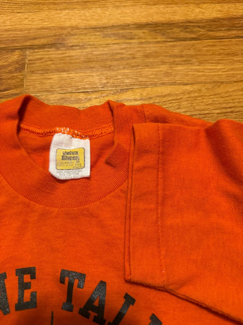 May include: Orange t-shirt with black lettering. The shirt has a crew neck and short sleeves. A tag is visible on the inside collar. The shirt is made of a blend of polyester and cotton. The text on the shirt is partially visible.