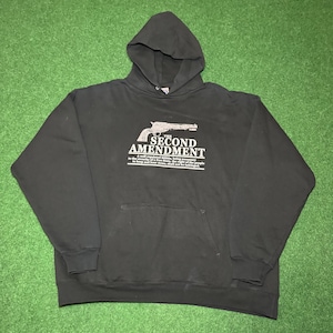 Könnte beinhalten: Schwarzer Kapuzenpullover mit einer Vordertasche und dem Text "SECOND AMENDMENT" über einer Pistolen-Grafik. Der Text darunter lautet: "Eine gut regulierte Miliz, die für die Sicherheit eines freien Staates notwendig ist, das Recht des Volkes, Waffen zu behalten und zu tragen, darf nicht beeinträchtigt werden."