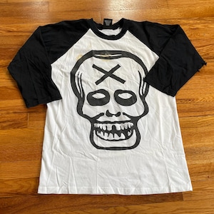 May include: A white and black baseball tee shirt with a skull graphic. The skull has a cross on its forehead and a dripping mouth.