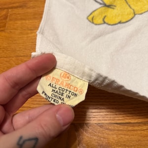 May include: A white cotton towel with a yellow tag that reads "FRANCO&reg; ALL COTTON MADE IN CHINA PRINTED IN". The towel features a cartoon character design in yellow and other colours.