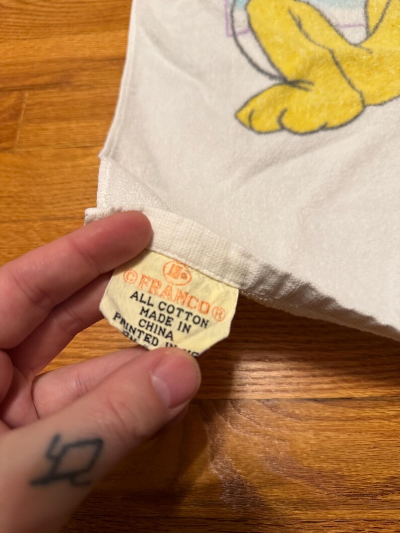 May include: A white cotton towel with a yellow tag that reads "FRANCO&reg; ALL COTTON MADE IN CHINA PRINTED IN". The towel features a cartoon character design in yellow and other colours.