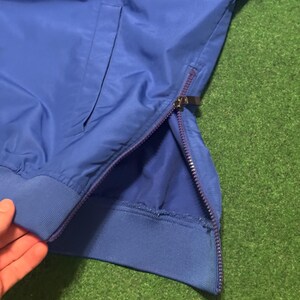 以下が含まれることがあります： ジッパーとポケットが付いた青いジャケットのクローズアップ。 ジャケットはリブ編みの袖口と、側面を走るジッパーが付いています。 ジャケットは滑らかで光沢のある素材で作られています。