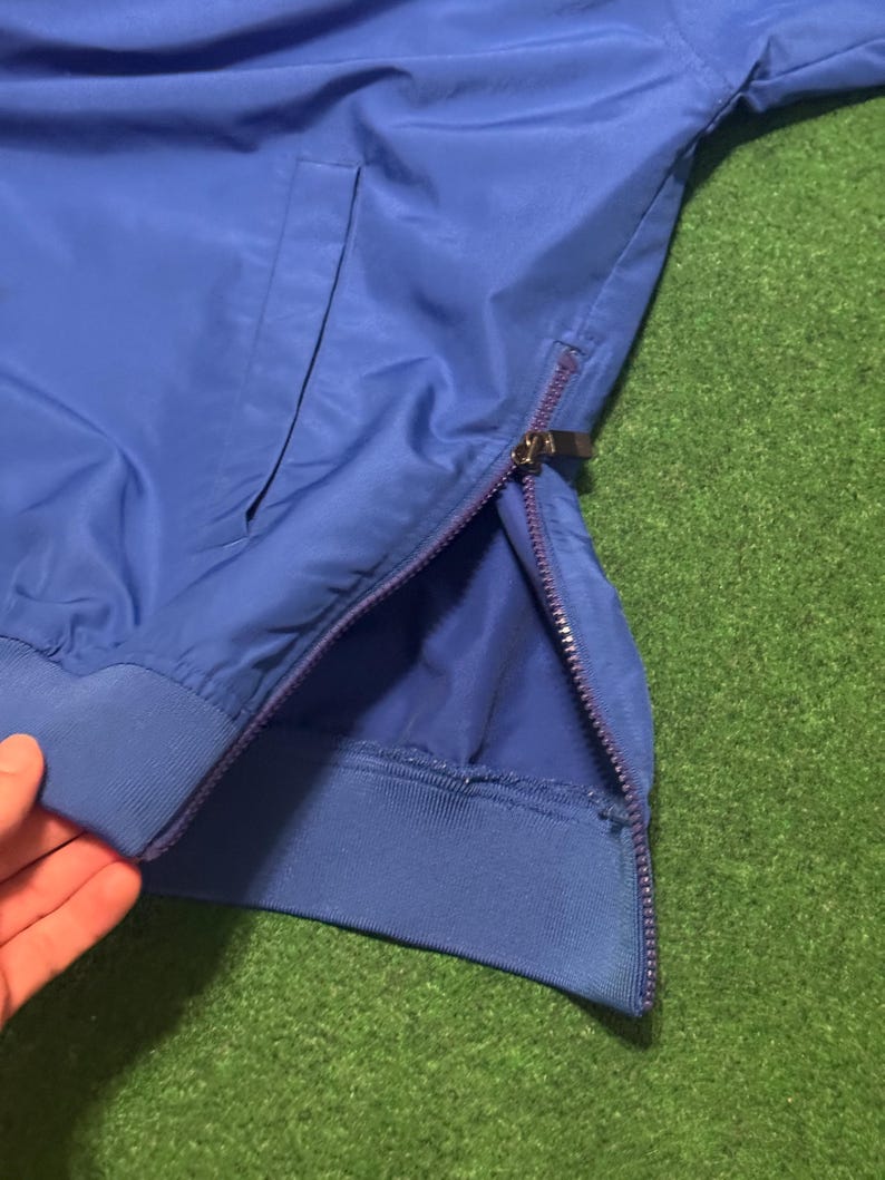 以下が含まれることがあります： ジッパーとポケットが付いた青いジャケットのクローズアップ。 ジャケットはリブ編みの袖口と、側面を走るジッパーが付いています。 ジャケットは滑らかで光沢のある素材で作られています。