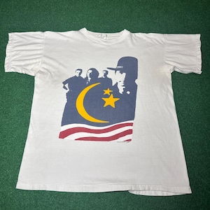 May include: A white short-sleeve t-shirt with ruffled sleeves. The front features a graphic print of three figures standing behind a large yellow crescent moon and two stars, with red and white stripes below and a silhouette of a person in a hat.