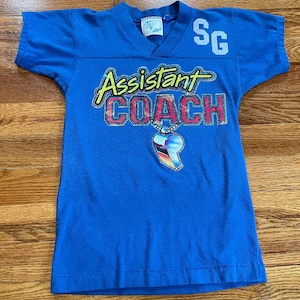 May include: A blue short-sleeved t-shirt with the text "Assistant Coach" in a colourful, glittery font. A whistle on a chain is depicted below the text. The number "SG" is printed on the upper right side of the shirt.