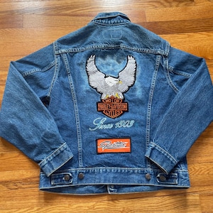 May include: A blue denim jacket with a white eagle and Harley Davidson logo patch on the back. The jacket also has a patch that says "Sportster" and the text "Since 1903" embroidered on the back.