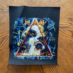 Può includere: Toppa in tessuto nero con un'immagine colorata di un viso che urla all'interno di un triangolo. Il testo "Def Leppard" e "Hysteria" è visibile.