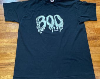 Peut inclure: T-shirt noir avec un motif blanc représentant le mot "BOO" qui coule avec de la bave. Le motif est de style vintage.