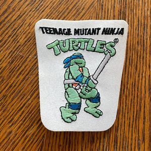 May include: A white embroidered patch featuring a Teenage Mutant Ninja Turtles logo with the text "Teenage Mutant Ninja Turtles" and "Turtles" in black. A green ninja turtle with a blue bandana is holding a silver sword.