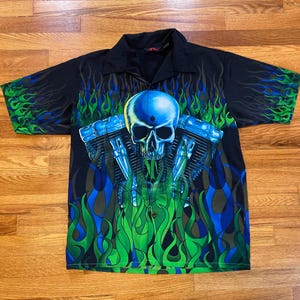 May include: A black short-sleeved button-up shirt with a blue and green skull and flames design. The skull is wearing a motorcycle helmet and is surrounded by flames.