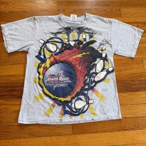 May include: A grey t-shirt with a bowling graphic. The design features a bowling ball, pins, and the text "USBC Youth Open Championships Indianapolis - 2009" in a circular emblem. The graphic has red, yellow, and blue accents.