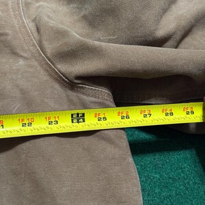 Puede incluir: Primer plano de una prenda marr&oacute;n con una cinta m&eacute;trica amarilla colocada encima. La cinta muestra medidas de 56 cm a 74 cm. La prenda parece estar hecha de un material duradero, posiblemente similar a la lona.