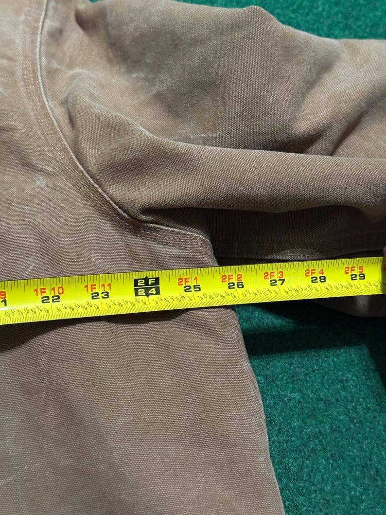 Puede incluir: Primer plano de una prenda marr&oacute;n con una cinta m&eacute;trica amarilla colocada encima. La cinta muestra medidas de 56 cm a 74 cm. La prenda parece estar hecha de un material duradero, posiblemente similar a la lona.