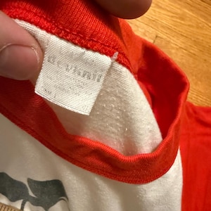 Pu&ograve; includere: Primo piano di una maglietta in stile baseball vintage con corpo bianco e maniche e colletto rossi. L'etichetta &egrave; visibile e un righello &egrave; in basso. La maglietta sembra usurata.