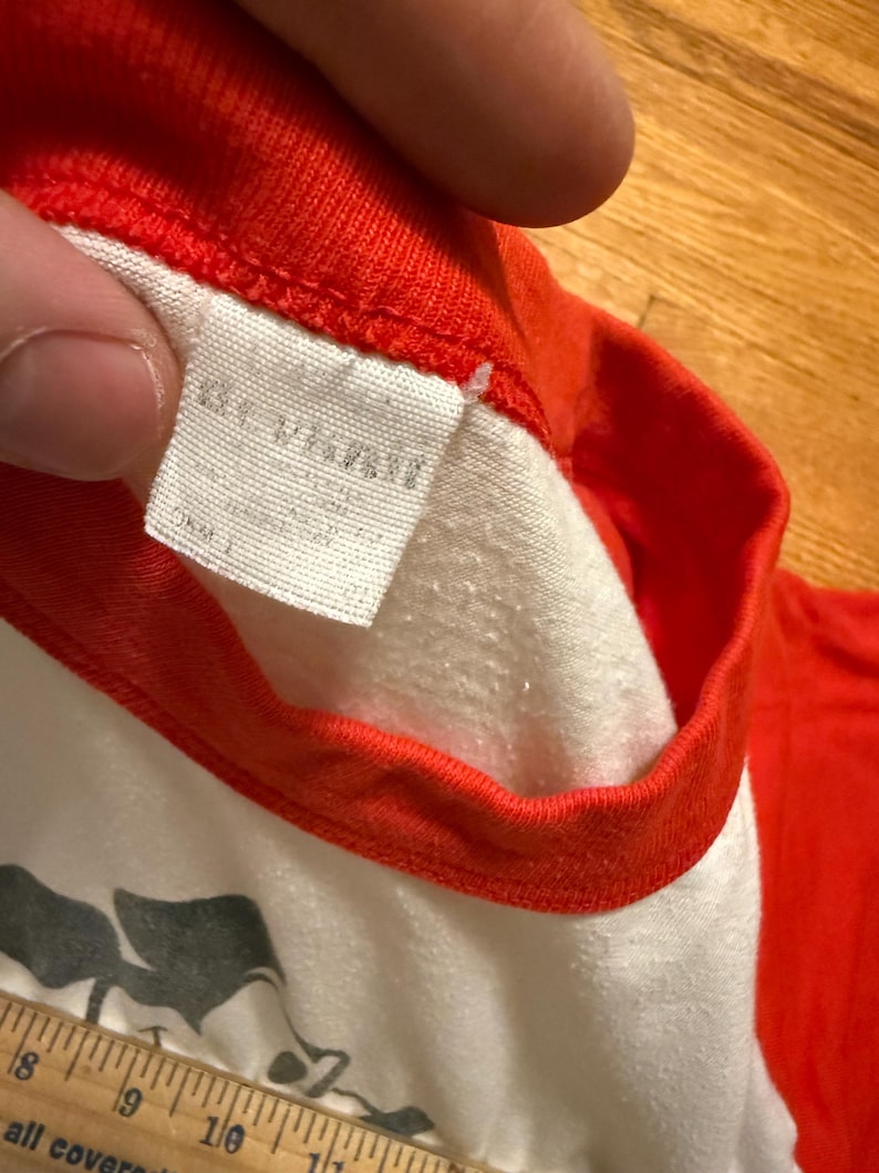 Pu&ograve; includere: Primo piano di una maglietta in stile baseball vintage con corpo bianco e maniche e colletto rossi. L'etichetta &egrave; visibile e un righello &egrave; in basso. La maglietta sembra usurata.