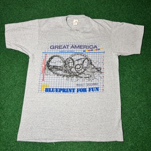 Puede incluir: Camiseta gris con un gráfico de un plano de una montaña rusa. El texto "Great America" y "Blueprint for Fun" está impreso en la camiseta.