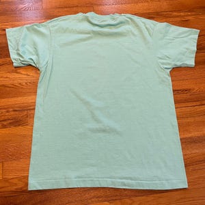 May include: A pale green, short-sleeved t-shirt. The shirt has a crew neck and is made of a soft material. The t-shirt is laid flat on a wooden surface. The shirt is a solid colour and has a simple, classic design.